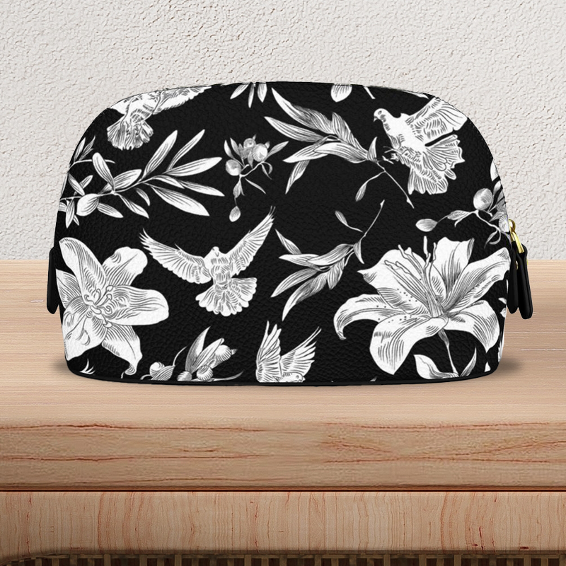 Back view view of a large, black, Nappa-textured leather makeup bag with gold-colored zipper and pulls, sits on a light-colored wooden console table. The bag is flat on the bottom with a curved top edge and has a bold white pattern of lilies, olive branches, and doves in an all-over print.