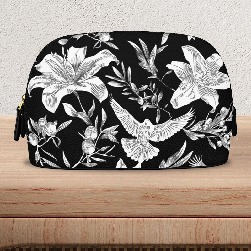 Frontal view view of a large, black, Nappa-textured leather makeup bag with gold-colored zipper and pulls, sits on a light-colored wooden console table. The bag is flat on the bottom with a curved top edge and has a bold white pattern of lilies, olive branches, and doves in an all-over print.