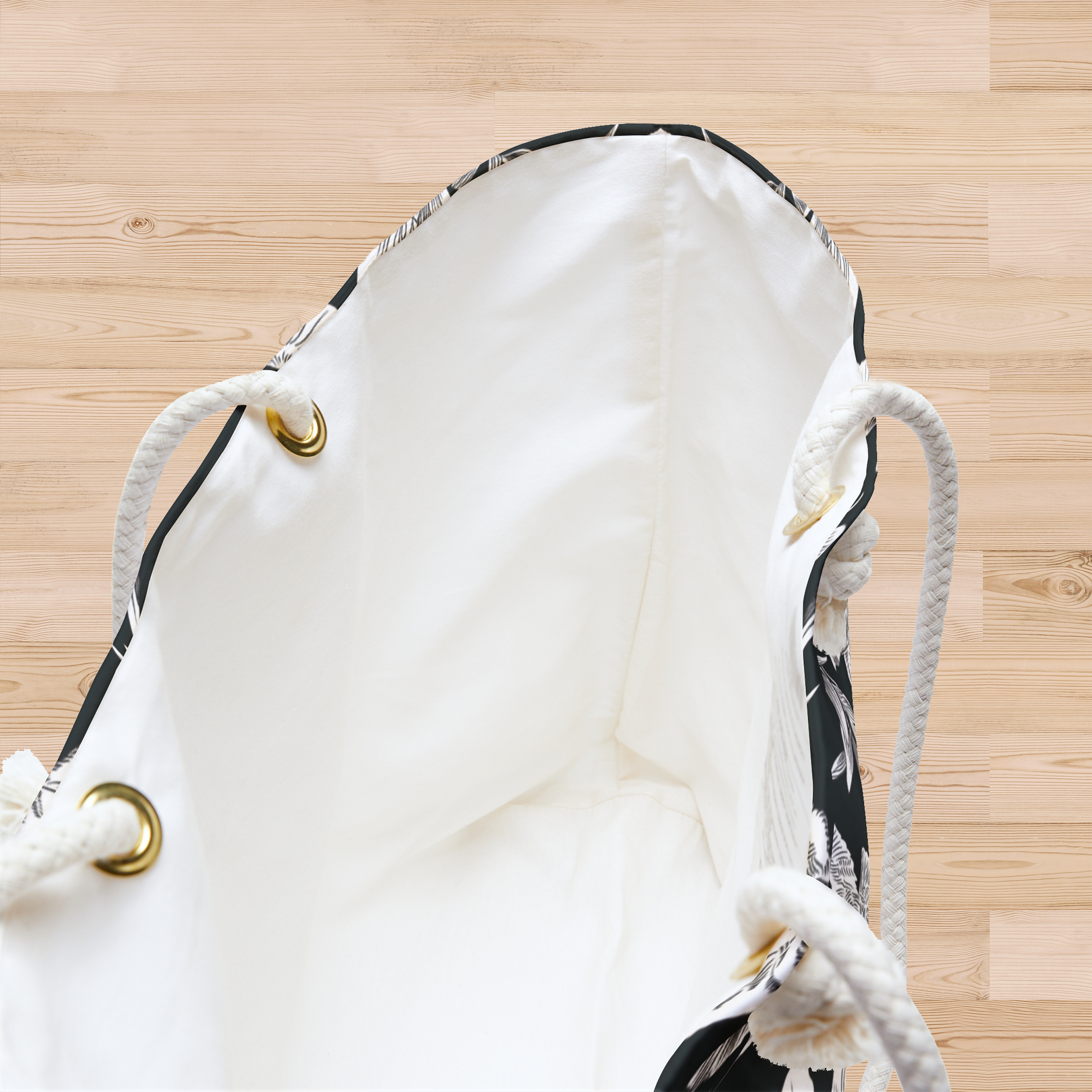 A close-up of the white interior of a large-capacity weekender tote with thick rope handles falling at either side, sits open on a pine, hardwood floor. A little bit of the bold white pattern of doves, lilies, and olive branches in an all-over printed pattern, can be seen along the edges and a partial side of the bag. Grommets for the rope handles are gold-colored.