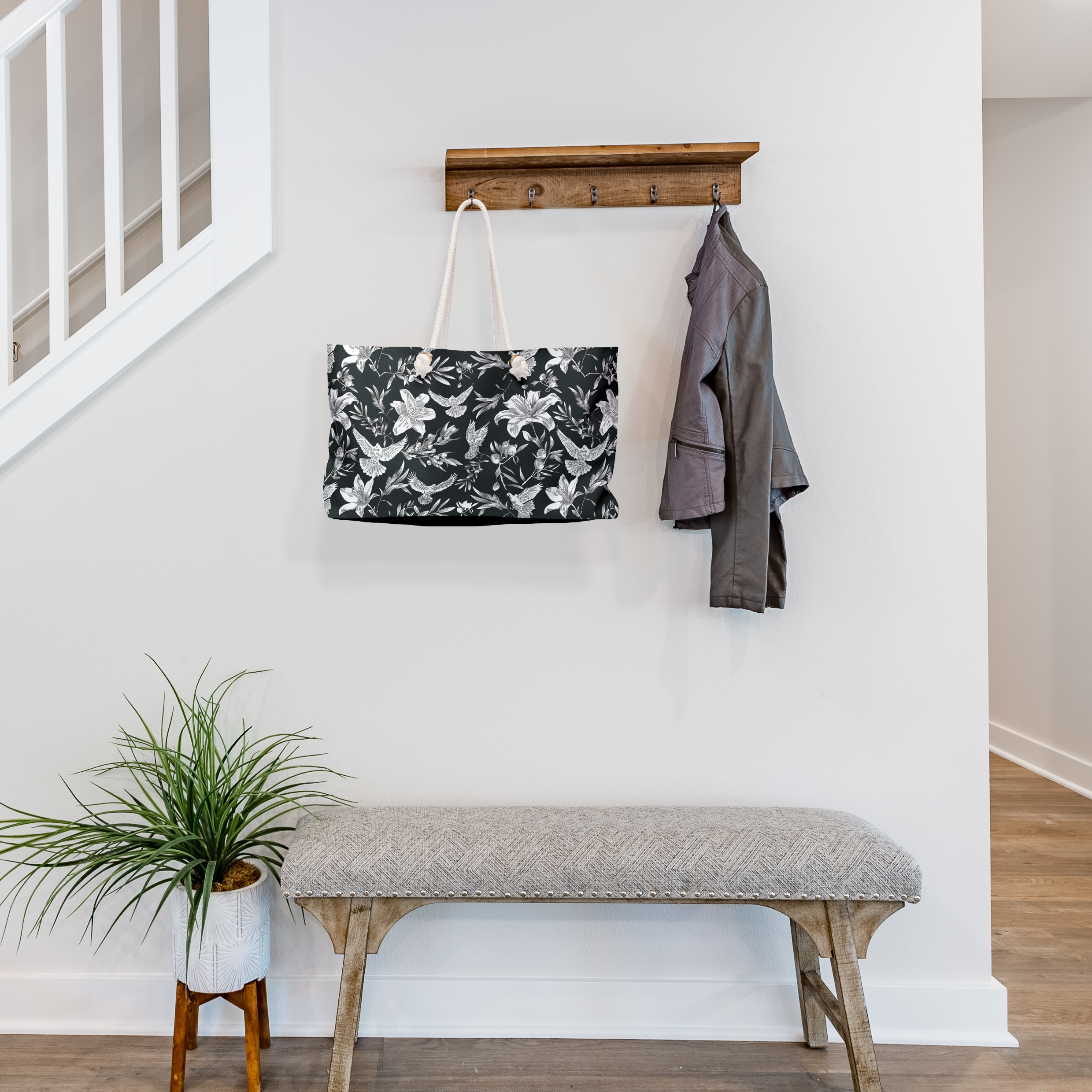 A large-capacity, black, weekender bag with  thick rope handles, hangs from a jacket rack in the entryway of a transitionally-styled house of neutral colors. The bag has a bold white pattern of doves, lilies, and olive branches in an all-over printed pattern. Grommets to accommodate the rope handles are gold-colored.