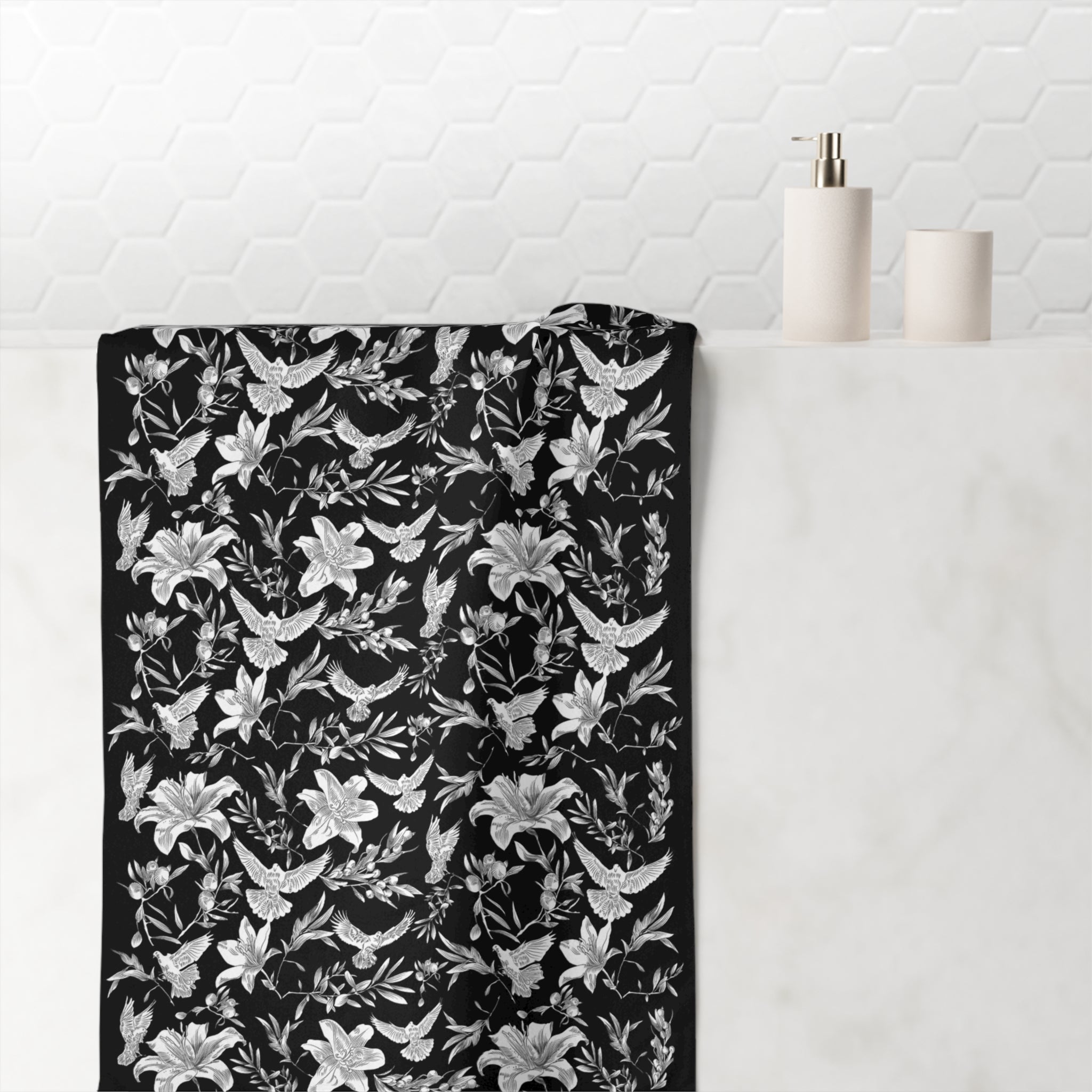 Black towel with white floral and dove pattern draped over a white surface with a white tiled wall background.