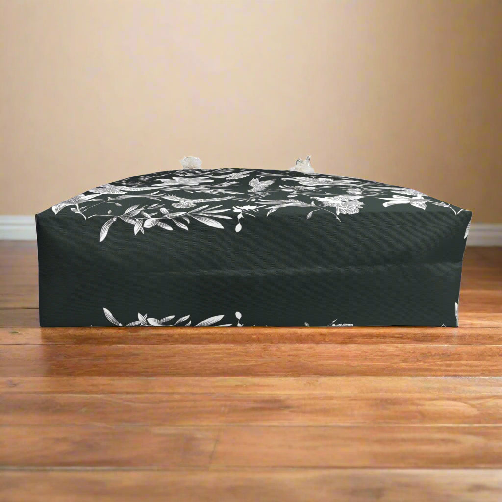 The base of a black tote bag with white floral pattern on a wooden floor against a beige wall.