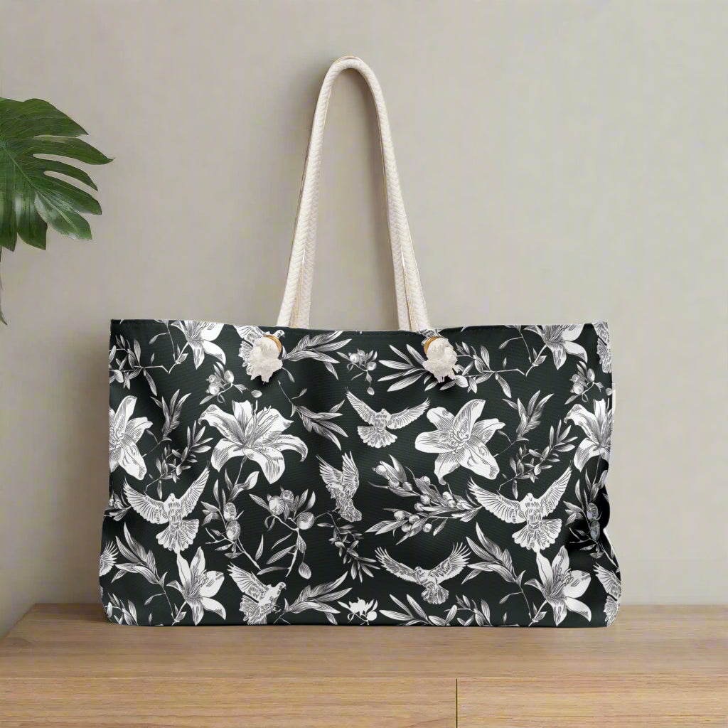 A large-capacity, black, weekender tote with a white, all-over pattern of doves, lilies, and olive branches with thick rope handles and gold-colored grommets stands against a white background.