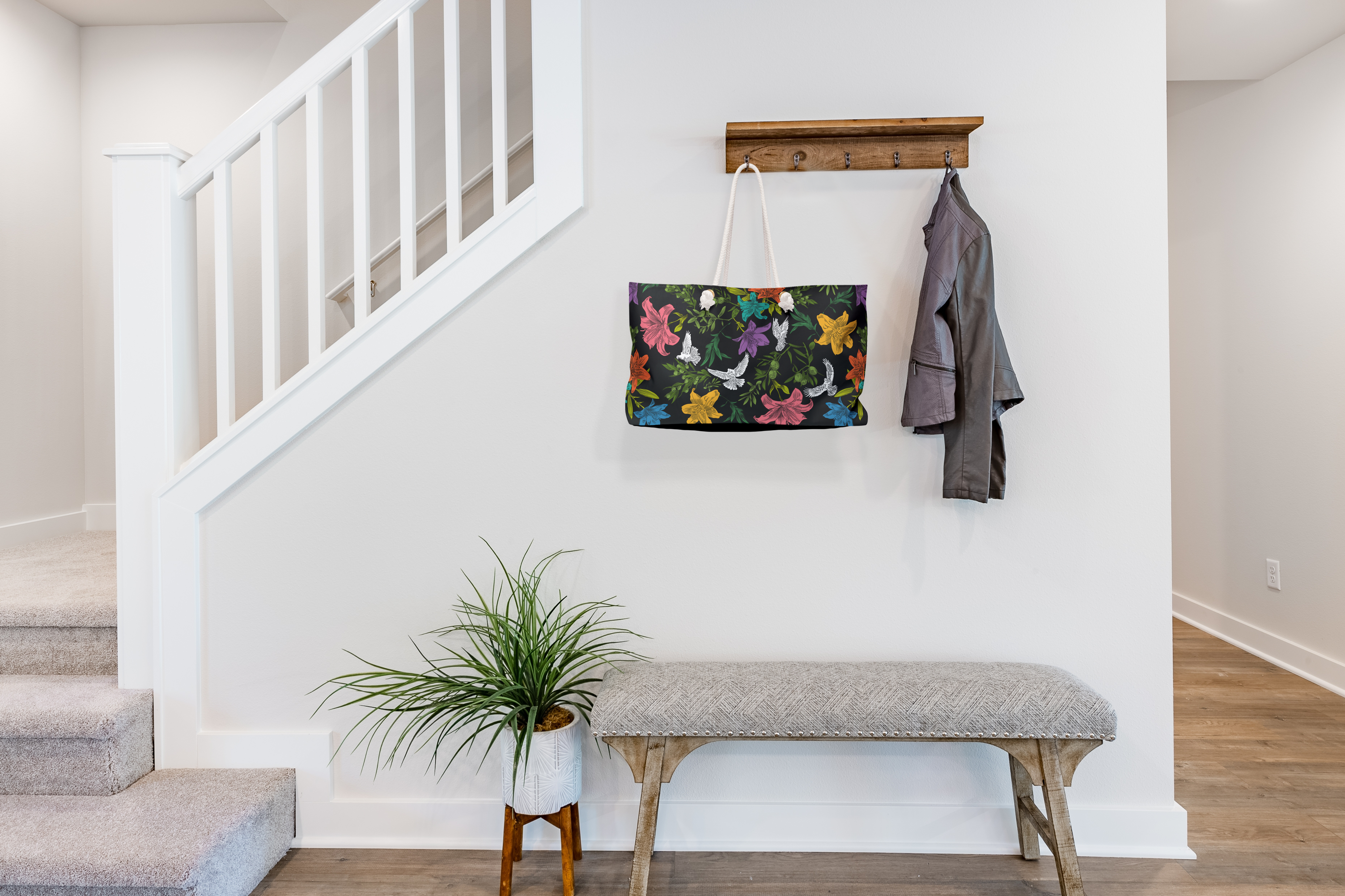 A large-capacity, black, weekender bag with thick rope handles, hangs from a jacket rack in the entryway of a transitionally-styled house of neutral colors. The bag has a bold, multi-colored pattern of doves, lilies, and olive branches in an all-over printed pattern. Grommets to accommodate the rope handles are gold-colored.