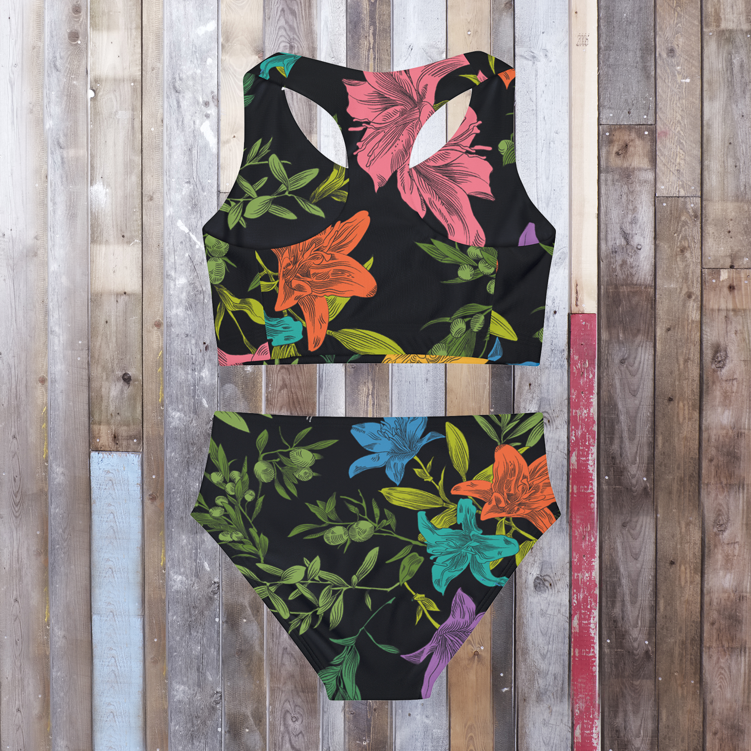 Back view of a two-piece, black, tank-style swimsuit laying flat on a hardwood floor, has an all-over, multi-colored pattern with flowers, olive branches, and doves. 