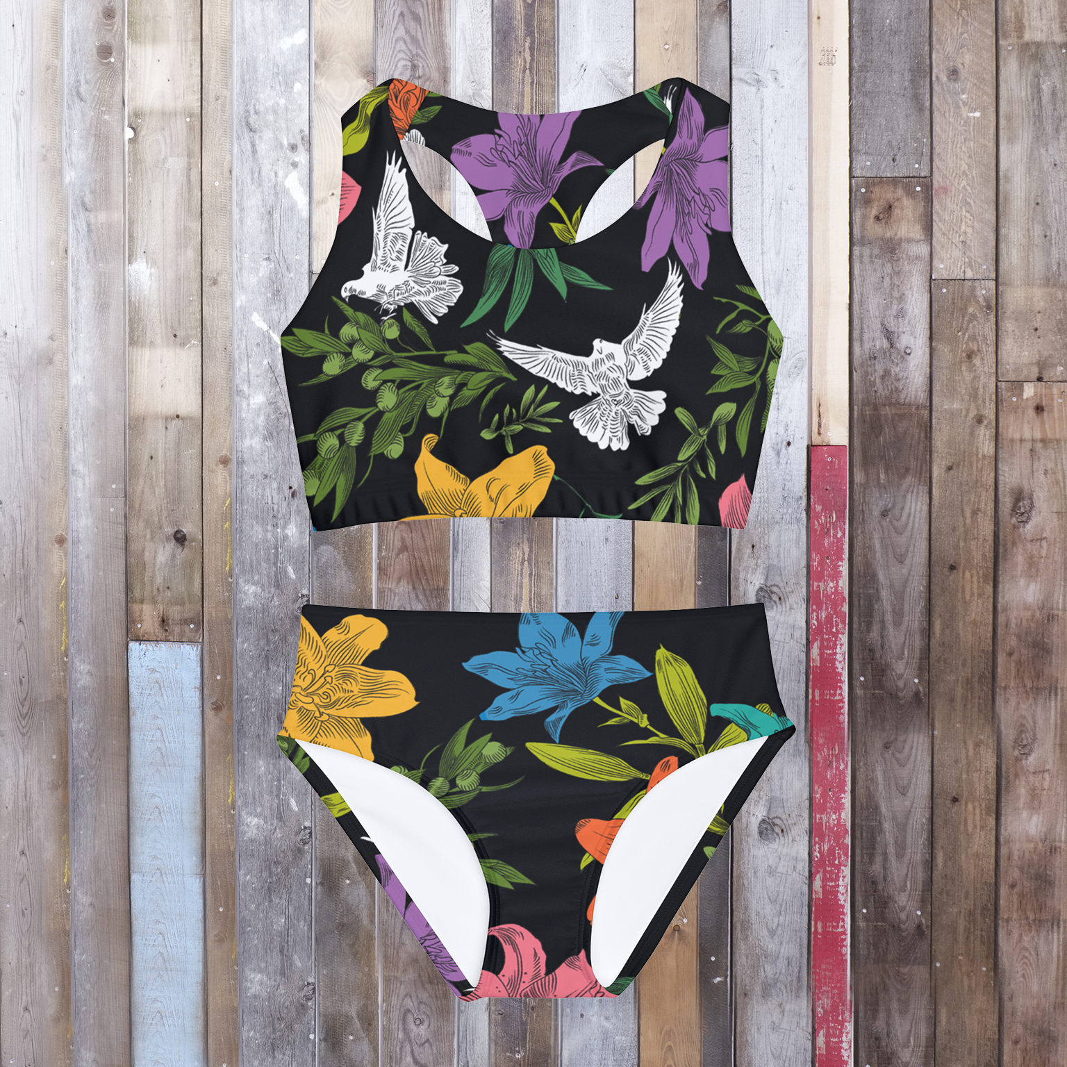 A two-piece, black tank-style swimsuit laying flat on a hardwood floor, has an all-over, multi-colored pattern with flowers, olive branches, and doves. 