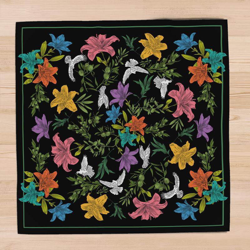 A square, black, silk scarf with green border and a bold, multi-colored floral pattern with white doves, lilies, and olive branches, lays atop a pine hardwood surface.