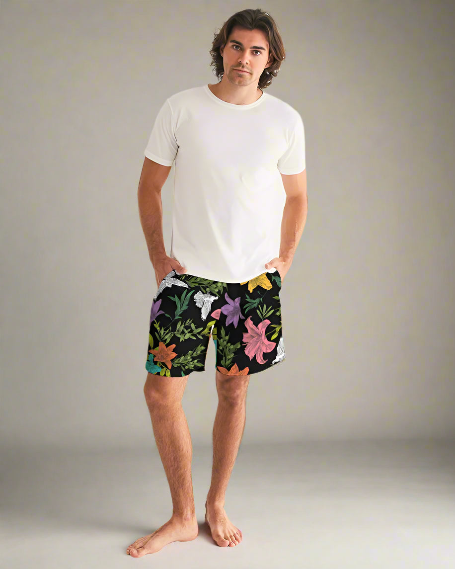 A man standing on a white background, wearing multicolored summer swim trunks with a geometric pattern.