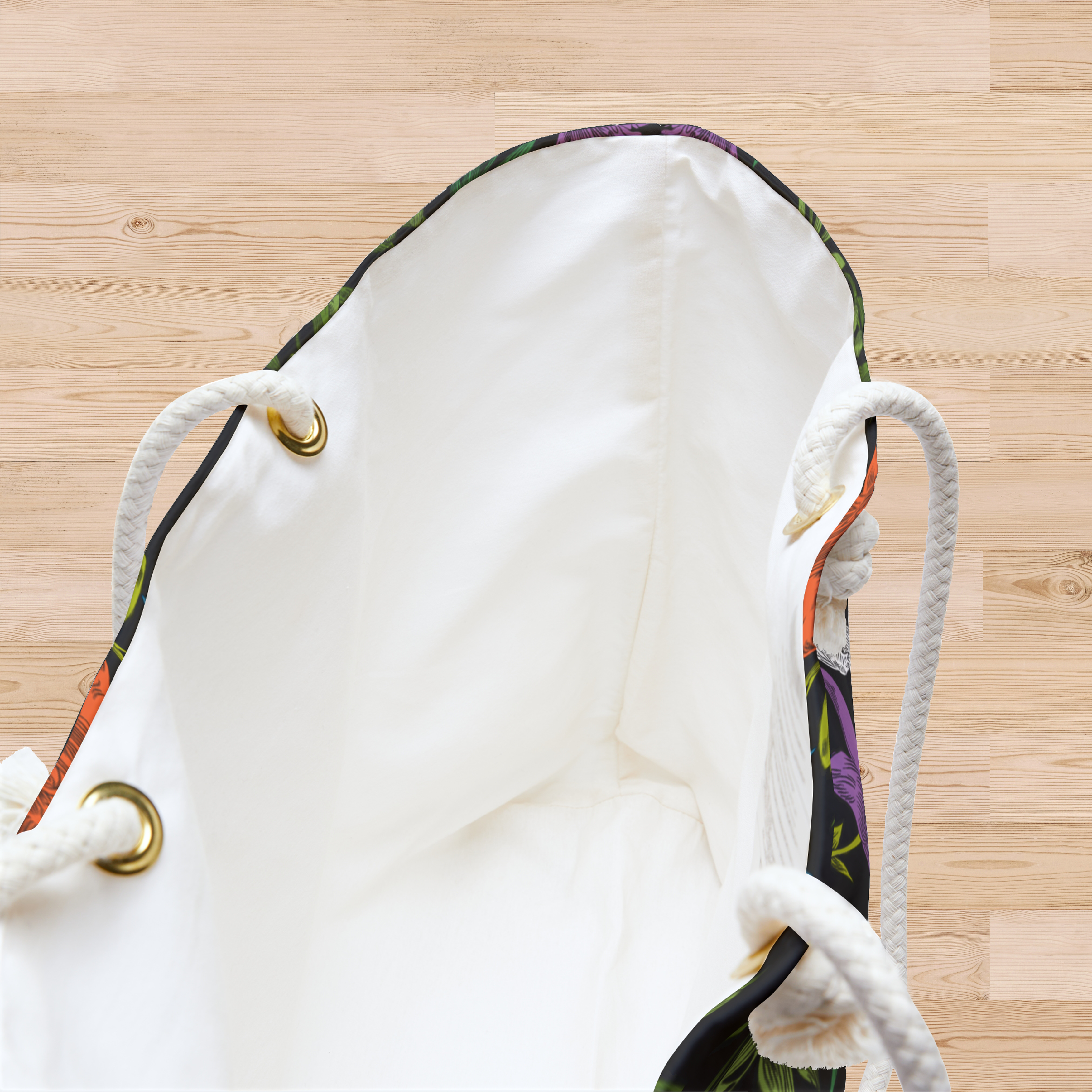 A close-up of the white interior of a large-capacity weekender tote with thick rope handles falling at either side, sits open on a pine, hardwood floor. A little bit of the bold, multi-colored pattern of doves, lilies, and olive branches in an all-over printed pattern, can be seen along the edges and a partial side of the bag. Grommets for the rope handles are gold-colored.