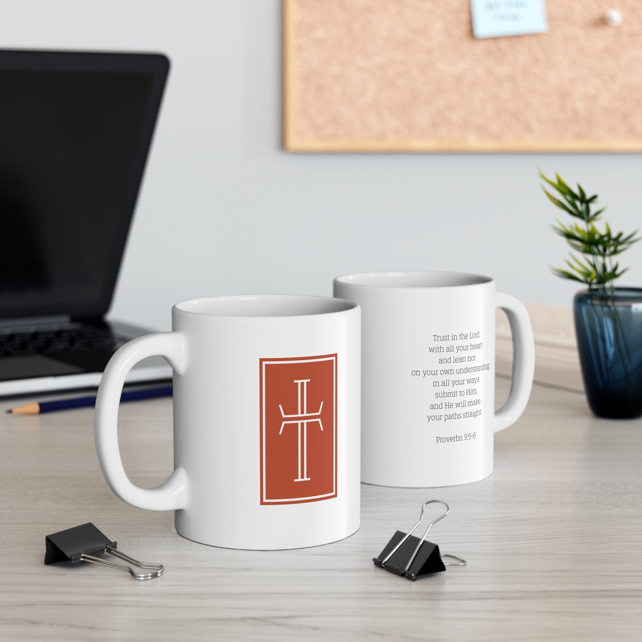 Two white mugs with a white cross against a warm, earthy-toned background on the front, and  the Biblical verse Proverbs 3:5-6 on the back. The mugs are sitting on a desk with office supplies.