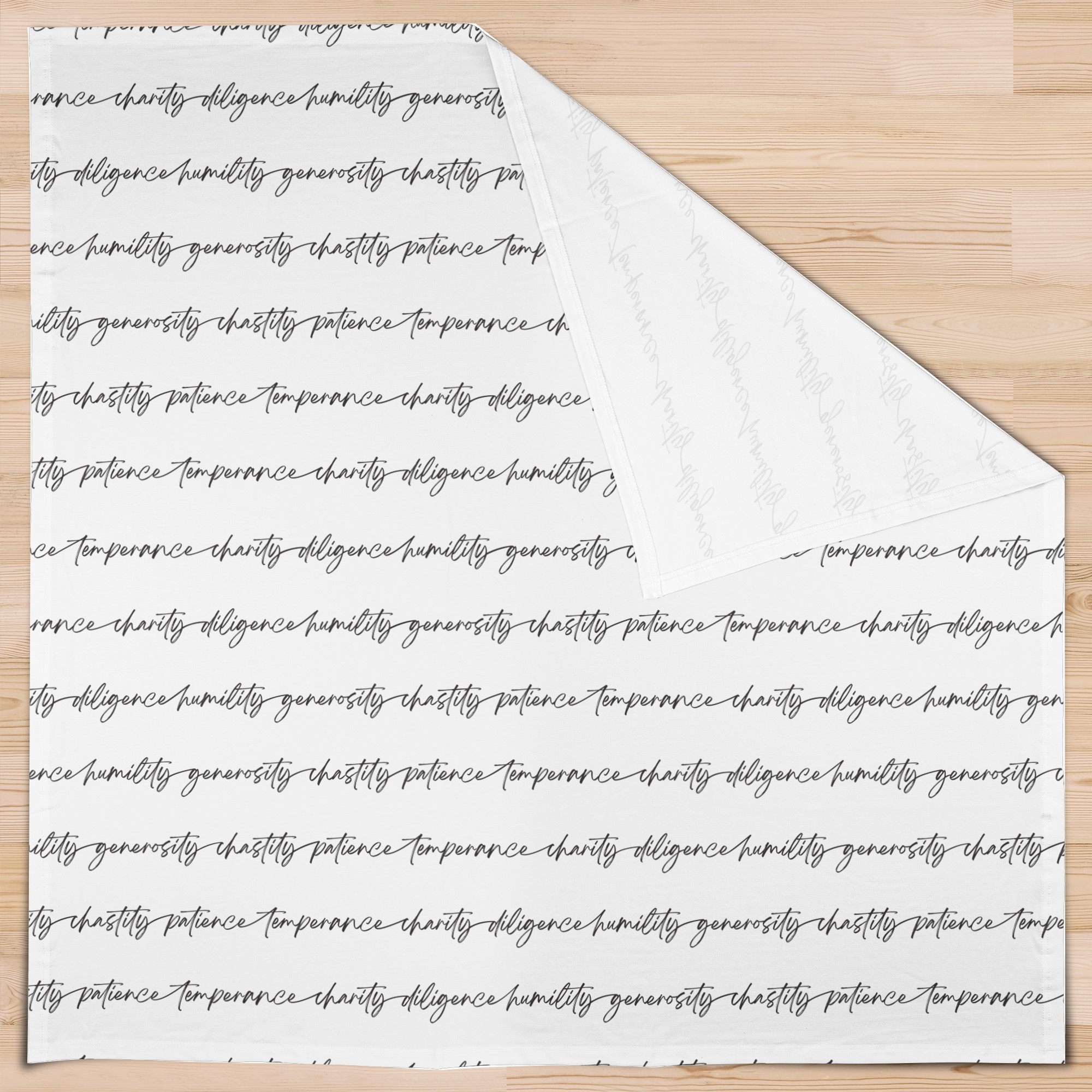 An overhead view of a white swaddle blanket with an all-over print of biblical virtues written in black script in continuous strips across the blanket face. One corner is folded back to display a white underside.