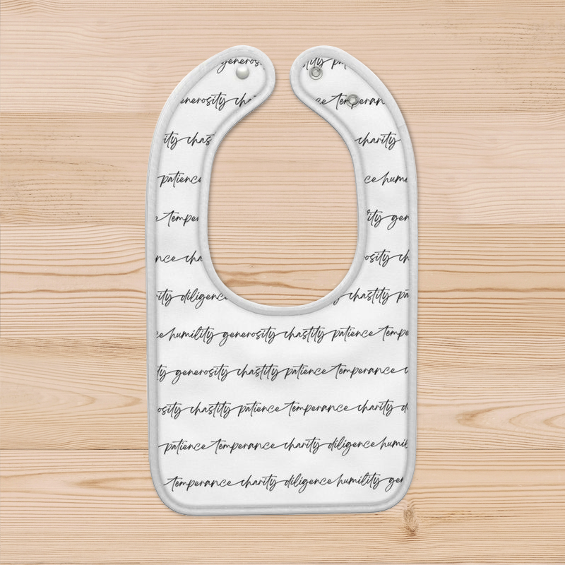 A white baby bib with biblical virtues written in black script in continuous strips across the garment. It has a white rolled border and two plastic snaps for alternate sizing options.