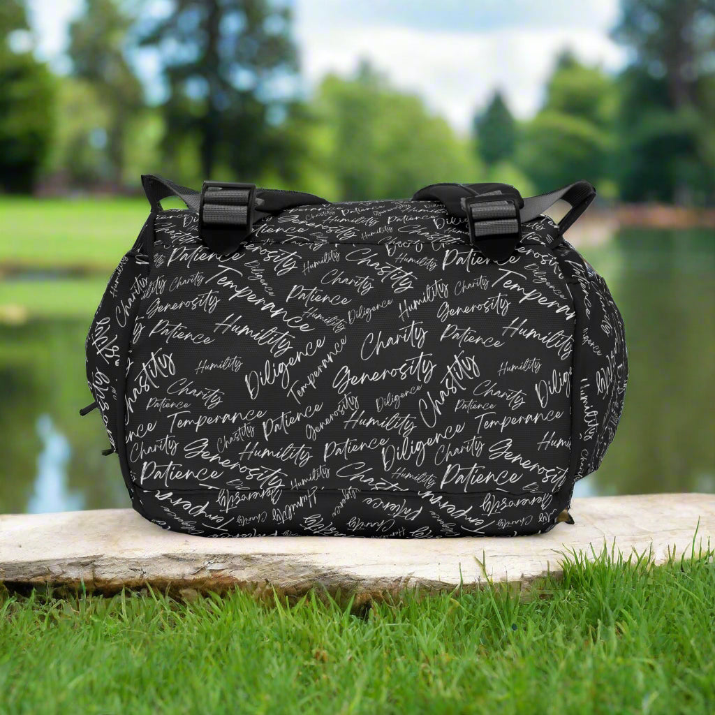 Bottom of backpack with adjustable straps with black and white scripted pattern laying on a flat stone by a lake and trees