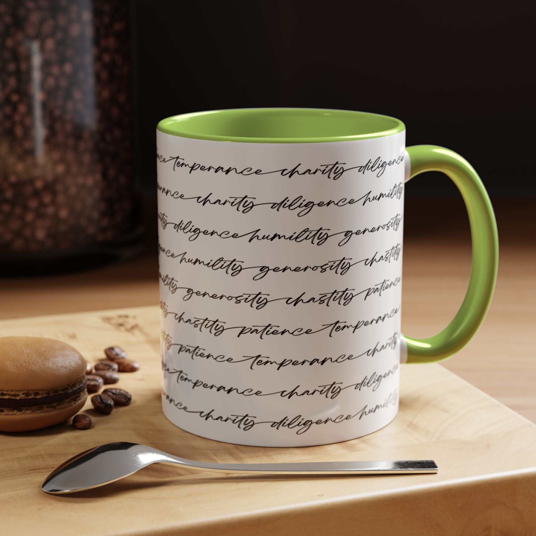 White mug with the seven virtues scripted along the outside, with green interior and handle on a wooden surface with coffee beans and a spoon.