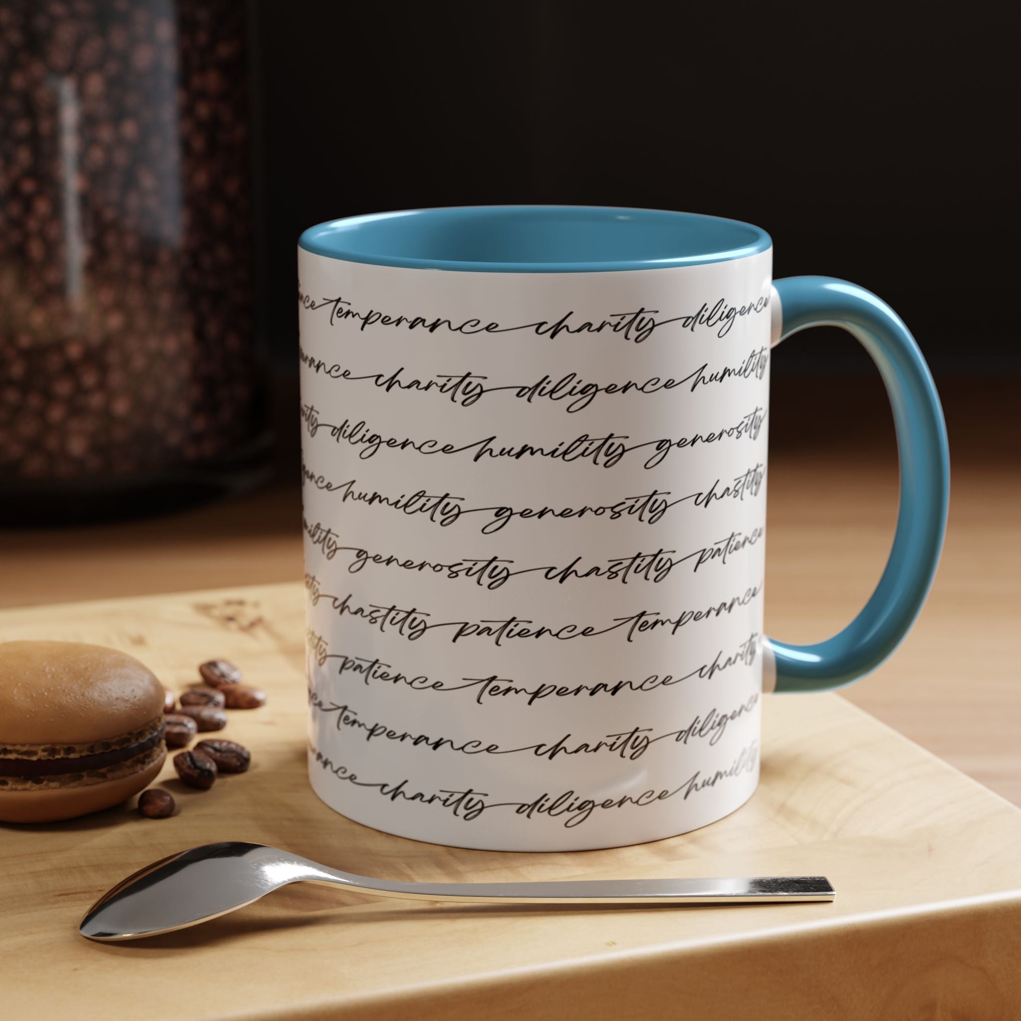 White mug with the seven virtues scripted along the outside, with blue interior and handle on a wooden surface with coffee beans and a spoon.