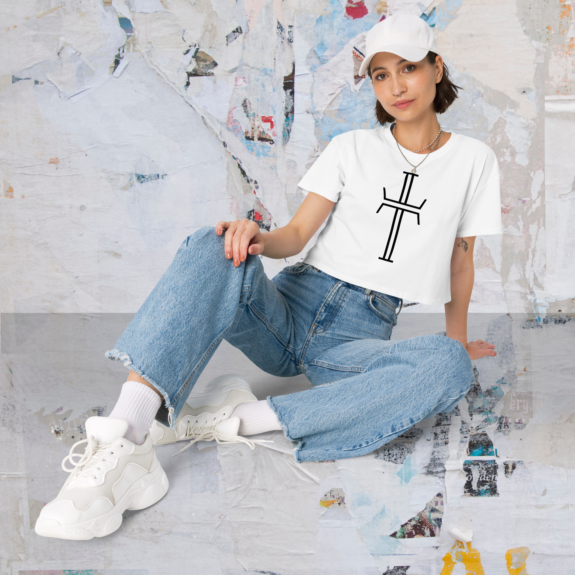 A female model is sitting on the ground in loose criss-cross style with her legs. She is wearing blue jeans and sneakers. Her white cropped tee shirt has a black Purpose logo on the front in a large bold graphic.