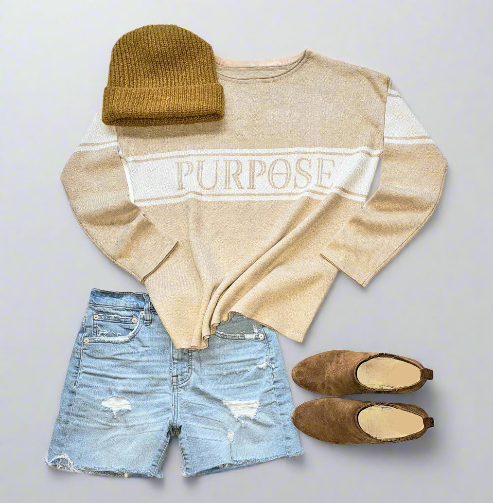 A beige, straight hemmed sweater with plain cuffs and rolled-neck detail is displayed against a white background. There are 3 white knitted stripes across the chest continuing to the upper arms. The middle, wide white stripe has the word "Purpose" written in beige capital letters across the center chest. A light brown beanie, brown suede ankle boots, and torn and faded denim shorts are arranged around the sweater.