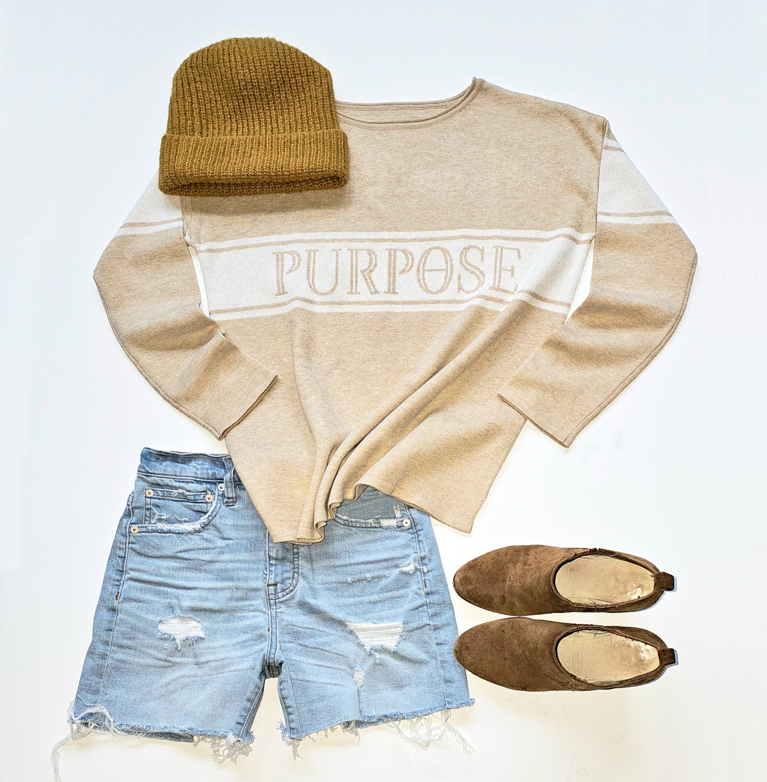 A beige, straight hemmed sweater with plain cuffs and rolled-neck detail is displayed against a white background. There are 3 white knitted stripes across the chest continuing to the upper arms. The middle, wide white stripe has the word "Purpose" written in beige capital letters across the center chest. A light brown beanie, brown suede ankle boots, and torn and faded denim shorts are arranged around the sweater.