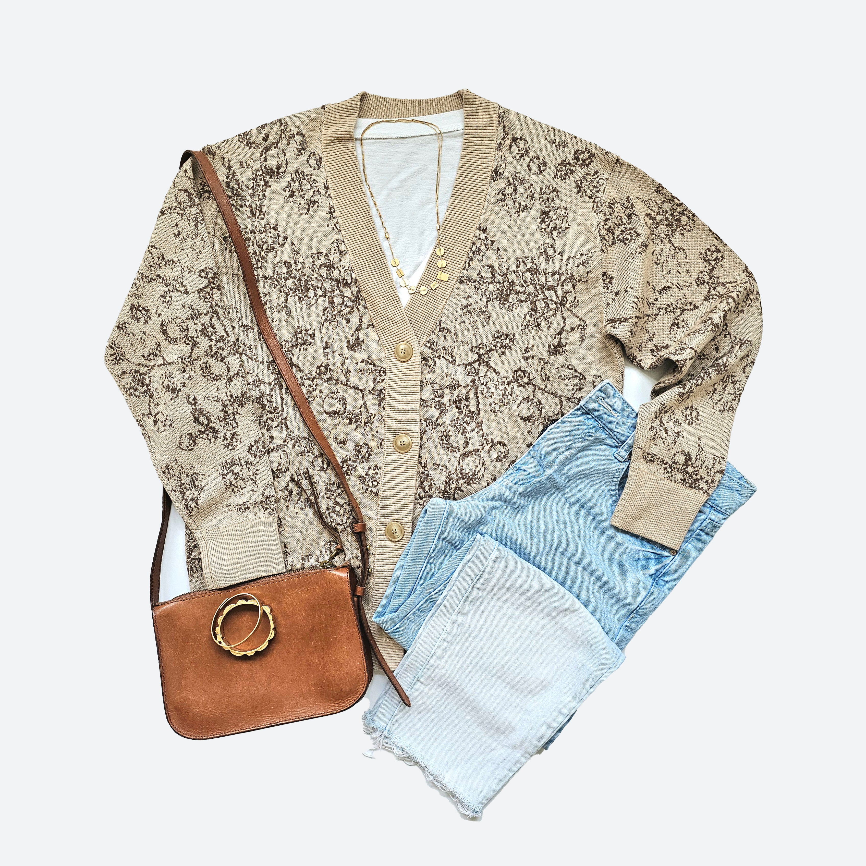 A beige knit, v-neck cardigan with large beige buttons is displayed against a white background. A brown floral pattern is knitted in an all-over pattern along the front. A pair of ombre-faded blue jeans, a small, camel colored shoulder bag, two gold-toned bangles and a gold-toned necklace are arranged around the cardigan. A white t-shirt peeks out from inside the sweater.