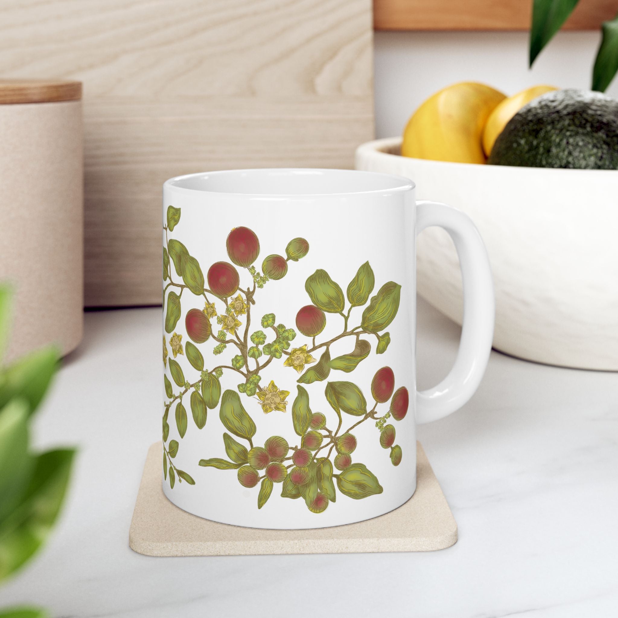 Left Side of white mug with floral and berry leaf design on a coaster, placed on a surface with a plant and bowl of fruit in the background.