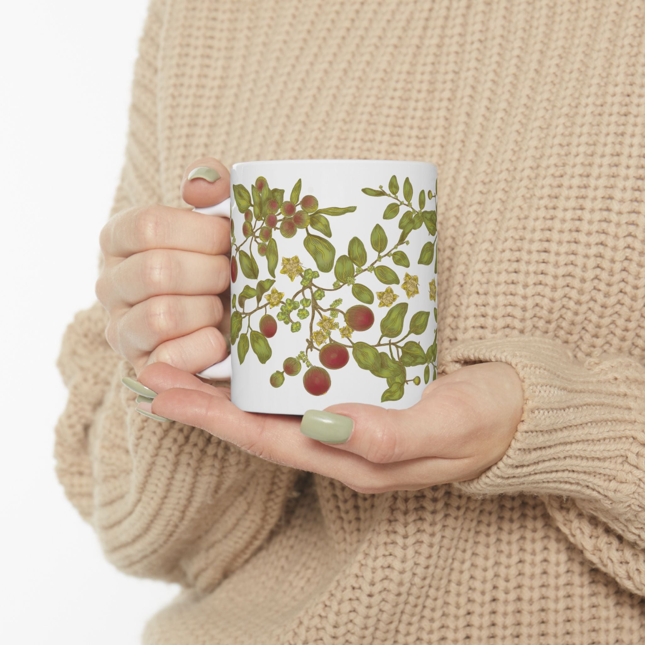 Woman in beige sweater holding a mug with a floral, berry, and leaf design against a neutral background.