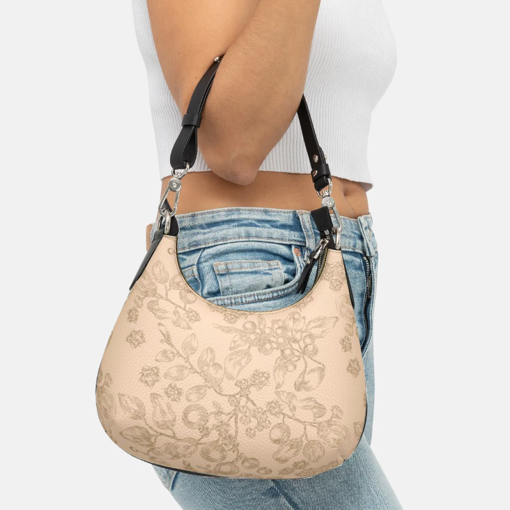 Close-up view of a slender woman's arm dangling a small, beige, leather purse from a bent elbow. The scoop-neck purse has a wider, bottom with rounded corners, and a dark brown line drawing of a plant all-over print pattern of leaves, berries, and flowers, with a black leather handle and gold-colored hardware. 