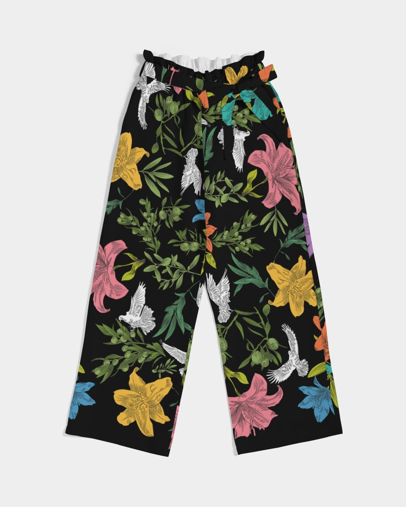 Front view of high-waisted, wide-leg black pants with a bold, multi-colored pattern of lilies, white doves, and olive branches in a large, all-over print with self-tie belt and elastic waist, laying flat against a white background.