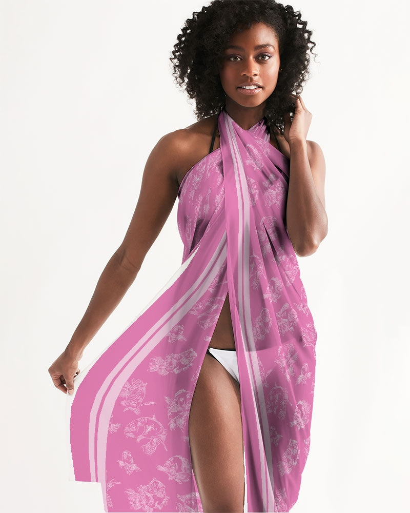 Closer view of a female model in a white string bikini wearing a pink, all-over printed, translucent, cover-up scarf worn as a full dress tied at her neck. The cover-up displays a white fish pattern and double white border along the edges.