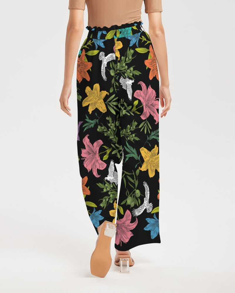 Back view of female model wearing high-waisted, wide-leg black pants with a bold, multi-colored pattern of lilies, white doves, and olive branches in a large, all-over print with self-tie belt and elastic waist.