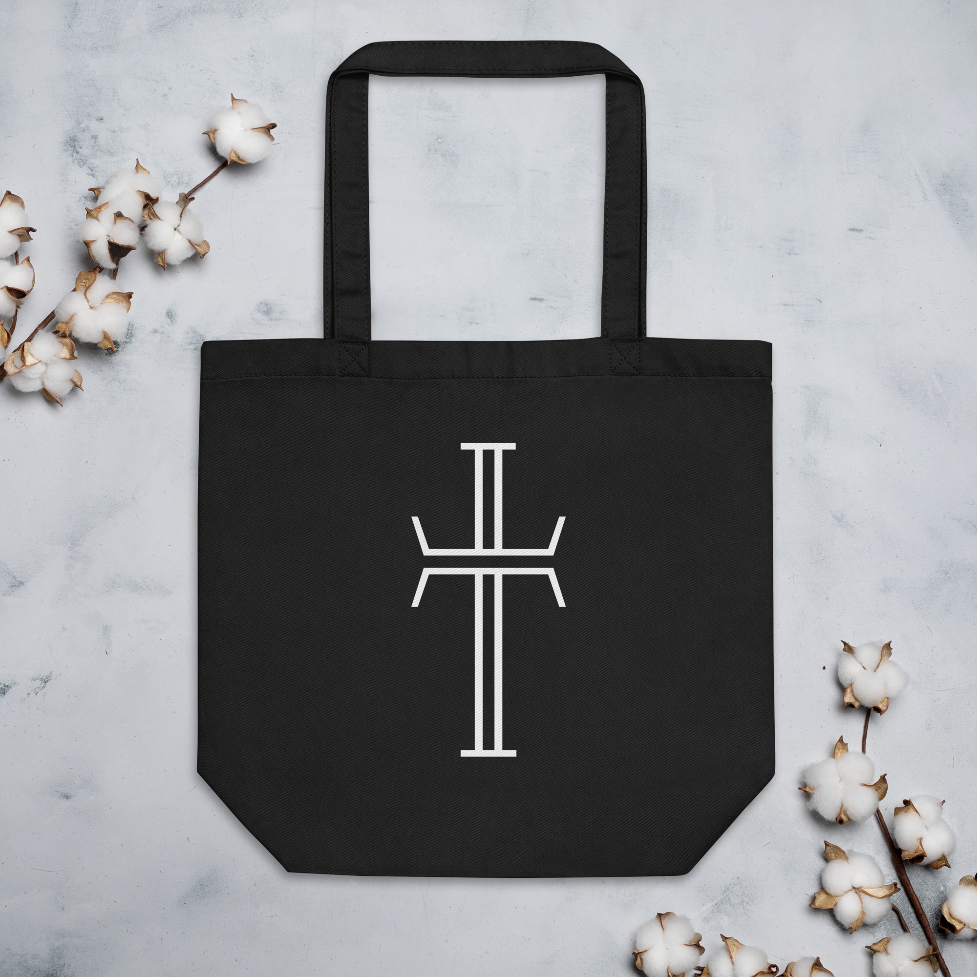 A black tote bag laying flat with the white Purpose logo cross on the front of the bag. The bag has long, generous handles.