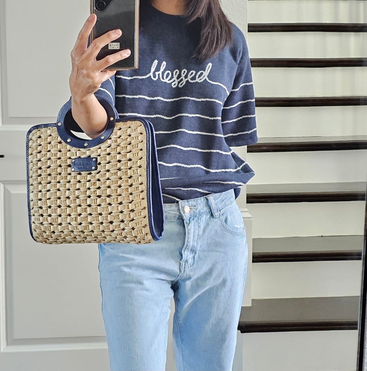 Person wearing a navy striped sweater with 'blessed' text, holding a woven handbag, standing on a staircase.
