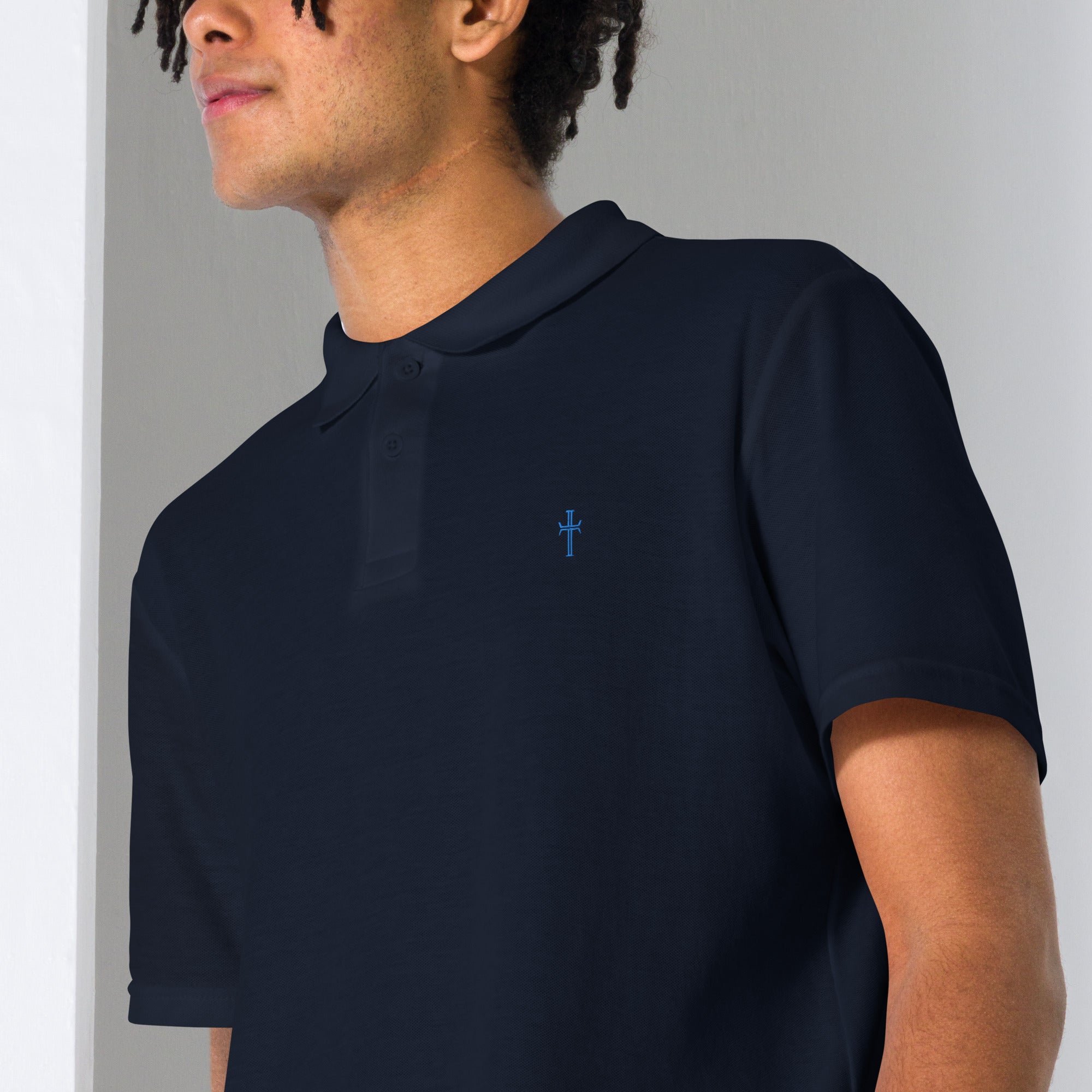 Close up of a male model wearing a navy pique knit polo with the Purpose cross logo in blue thread embroidered on the left chest area.